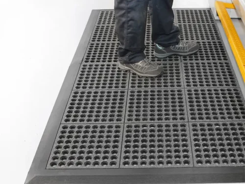 Industrijski otirači Steep – radnik stoji na crnom modularnom otiraču sa crnim ivicama uz industrijsku mašinu